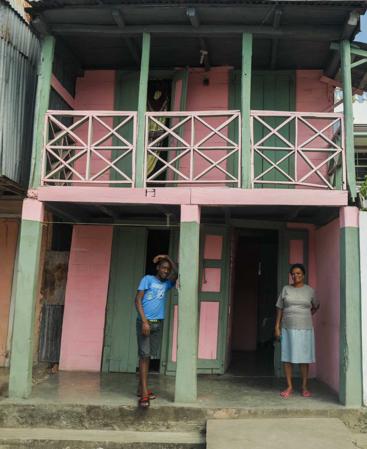 Two people stand smiling in front of a pink and green painted wooden house in Baradères, Haiti