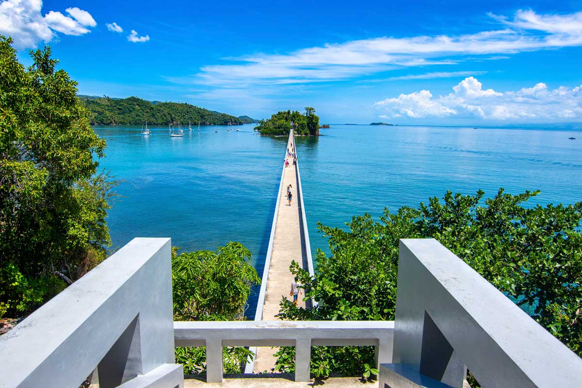 The long and narrow Samana Bay Bridge in the Dominican Republic stretching across the Atlantic ocean - connecting to smaller islets on a sunny day, as sailboats pass by along the calm water.